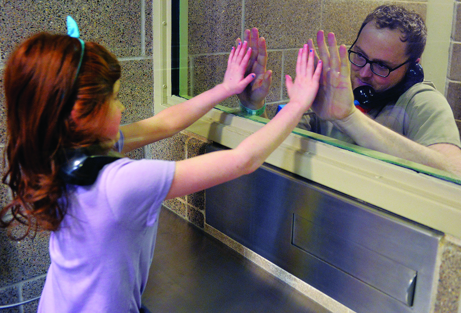 a father and daughter on opposite sides of plexiglass divider in jail